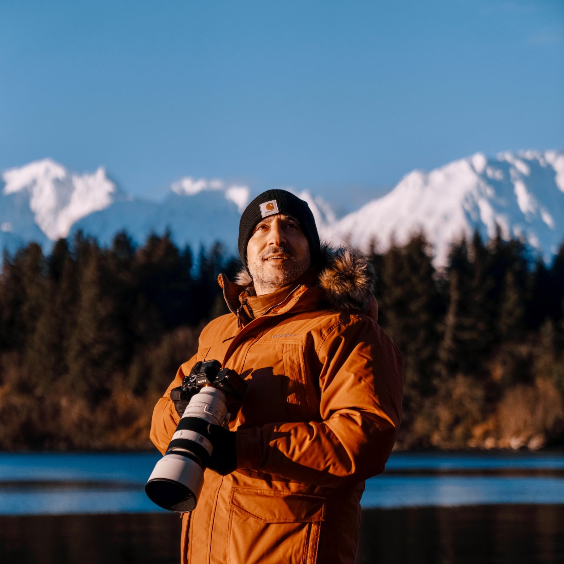 Hasan Akbas holding a camera in front of snow-covered Alaskan mountains at sunset