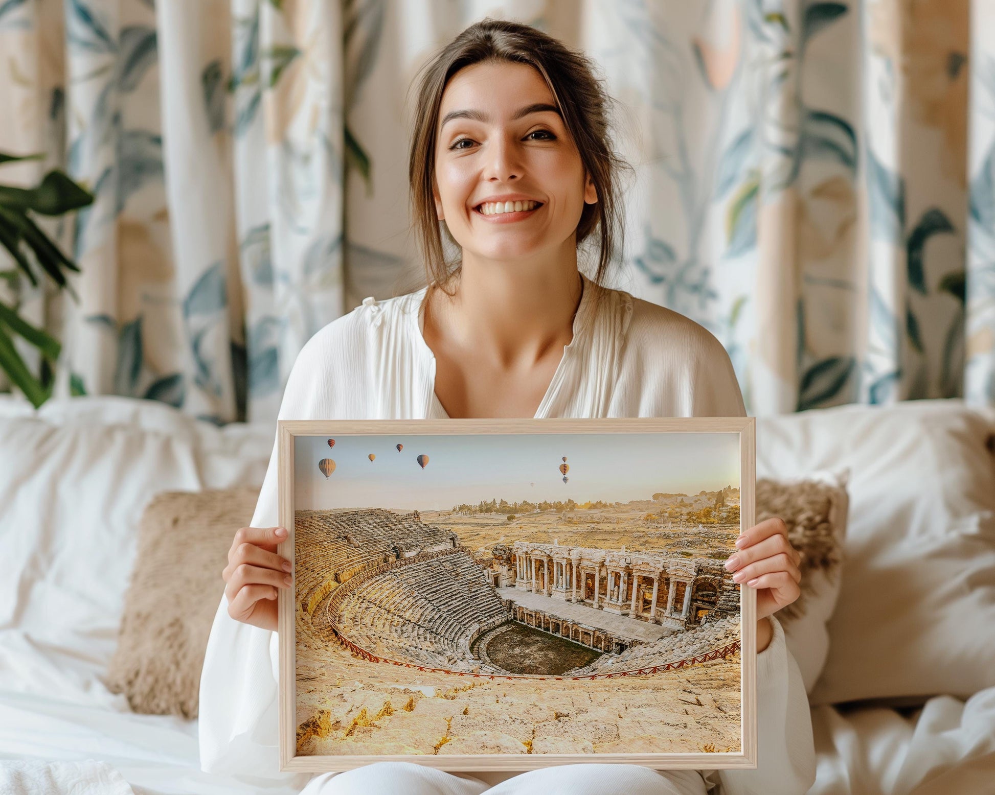 Smiling woman holding a light wood–framed print of Hierapolis ruins at sunrise, showing the ancient amphitheater and balloons in the sky in a cozy bedroom setting.