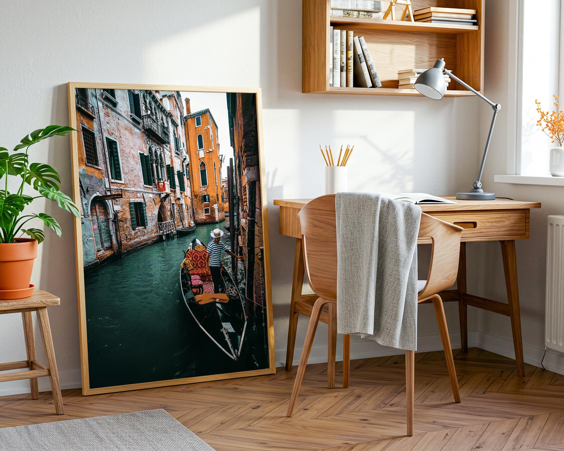 Large framed Venice canal photograph leaning beside a wooden desk and chair in a bright home office with plants and natural light.