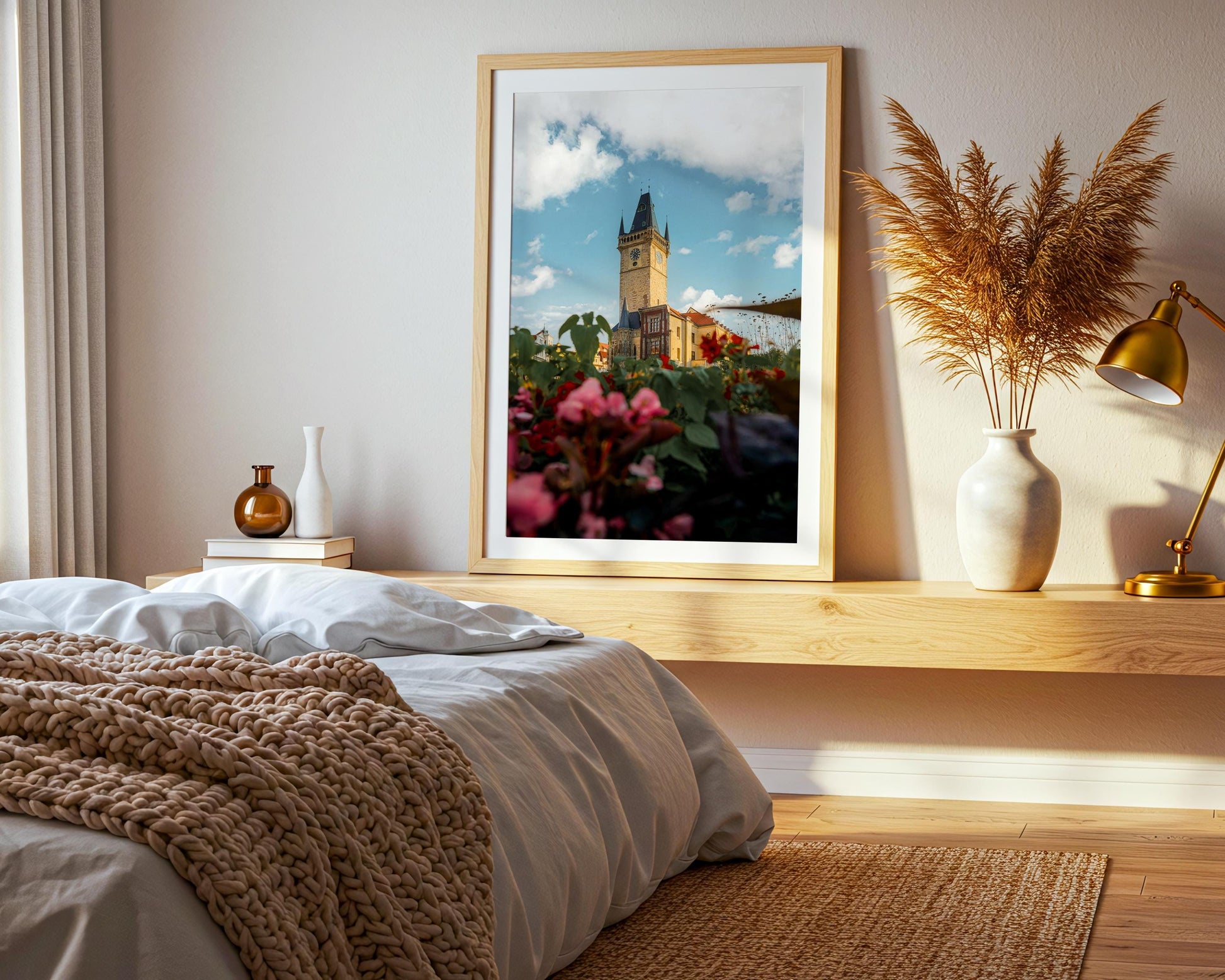 Large wood-framed Prague clock tower print leaning against a bedroom wall beside a bed, with soft morning light and neutral decor.