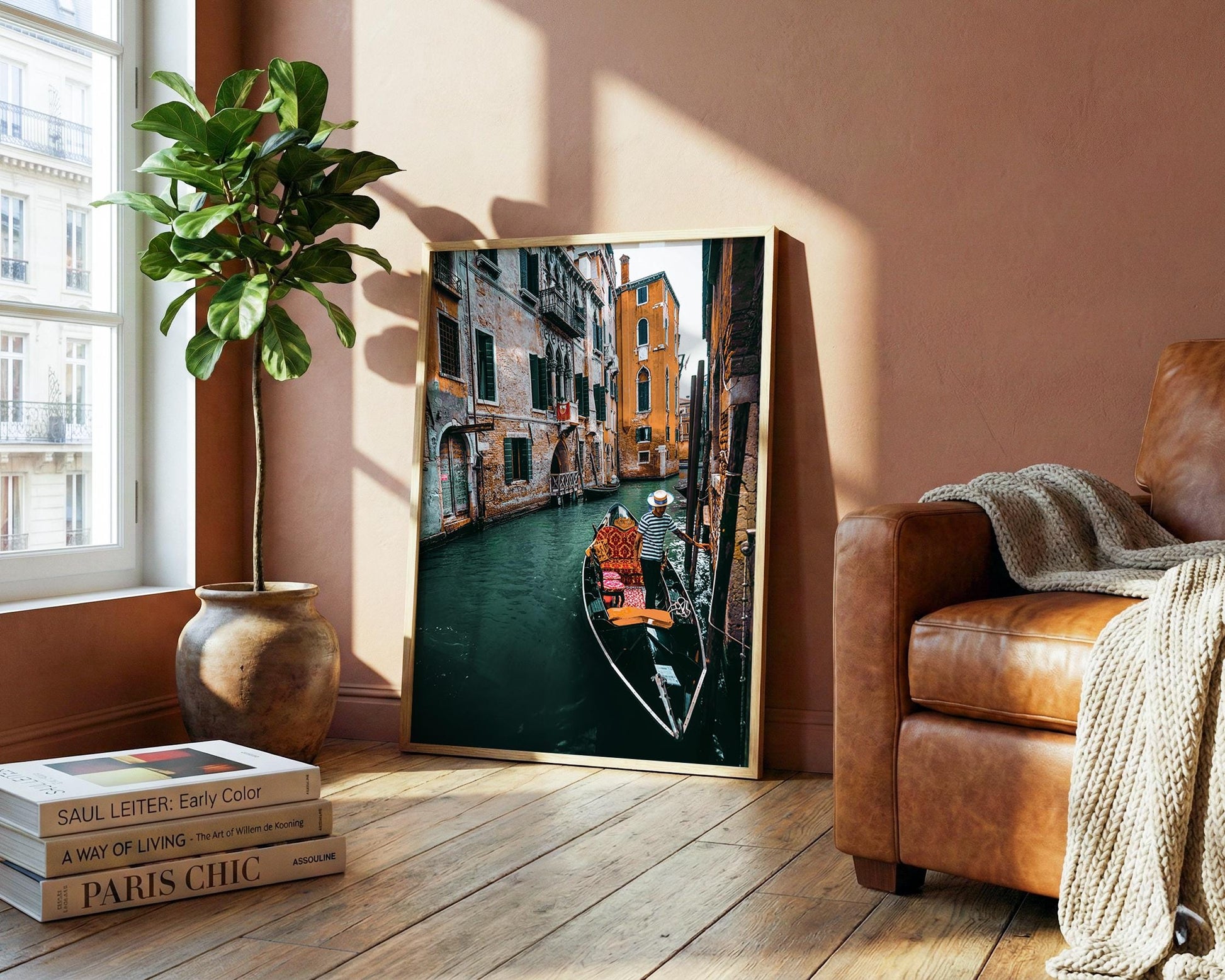 Framed Venice canal photograph leaning against a wall in a sunlit living room with a potted plant and a leather sofa.