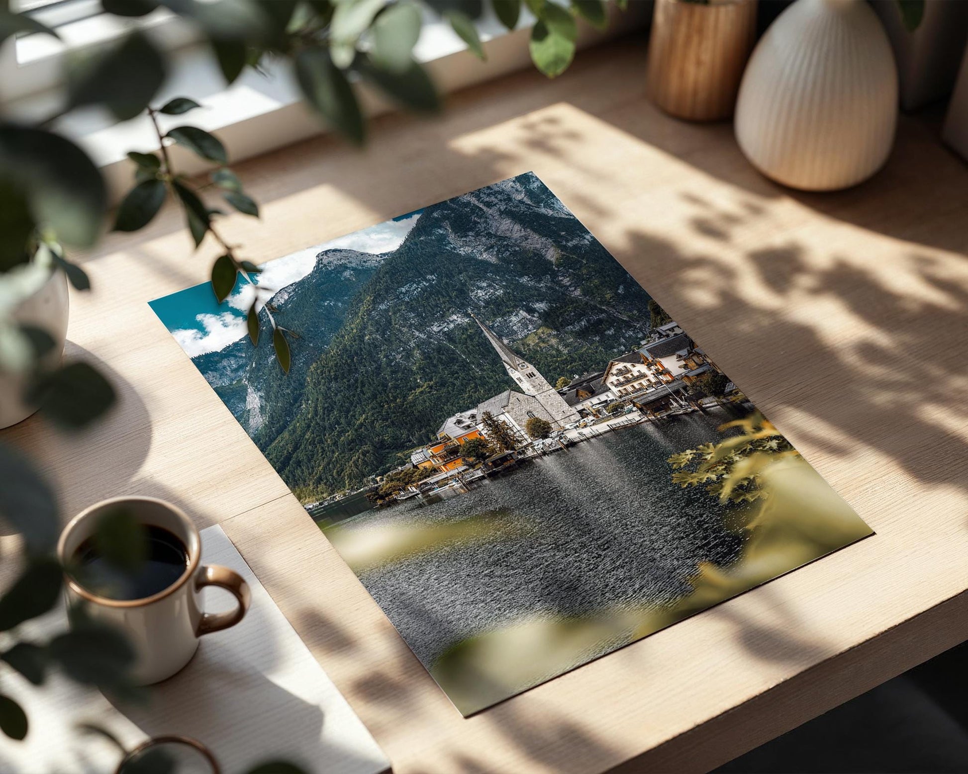Unframed Hallstatt village photo fine art print lying on a wooden table beside a coffee mug, with leafy shadows and warm window light.