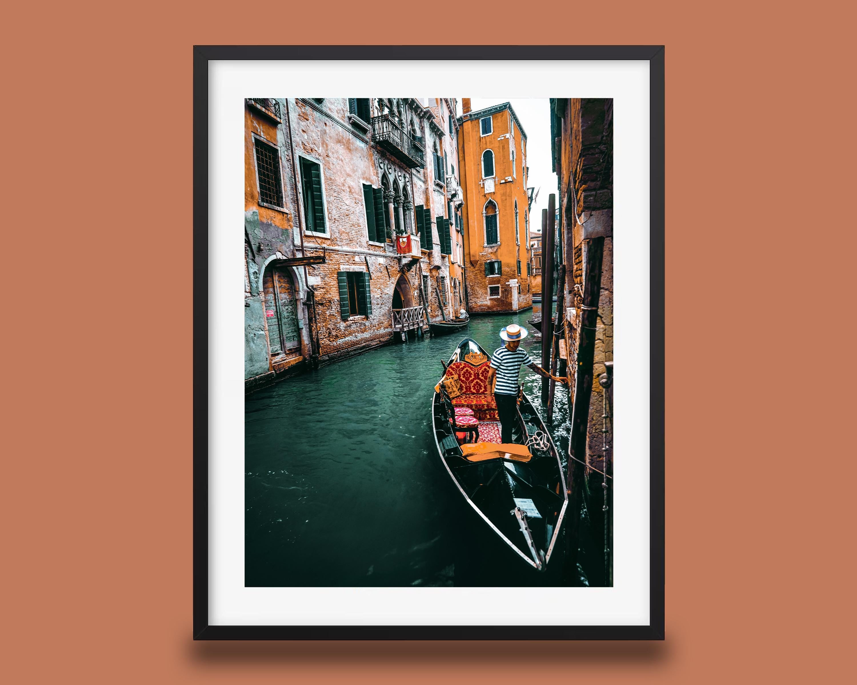 Black-framed print with a white mat showing a Venice canal scene, a gondolier standing in a gondola beside weathered brick buildings with green shutters and Warm Terracotta, Burnt Orange walls, over deep green water.